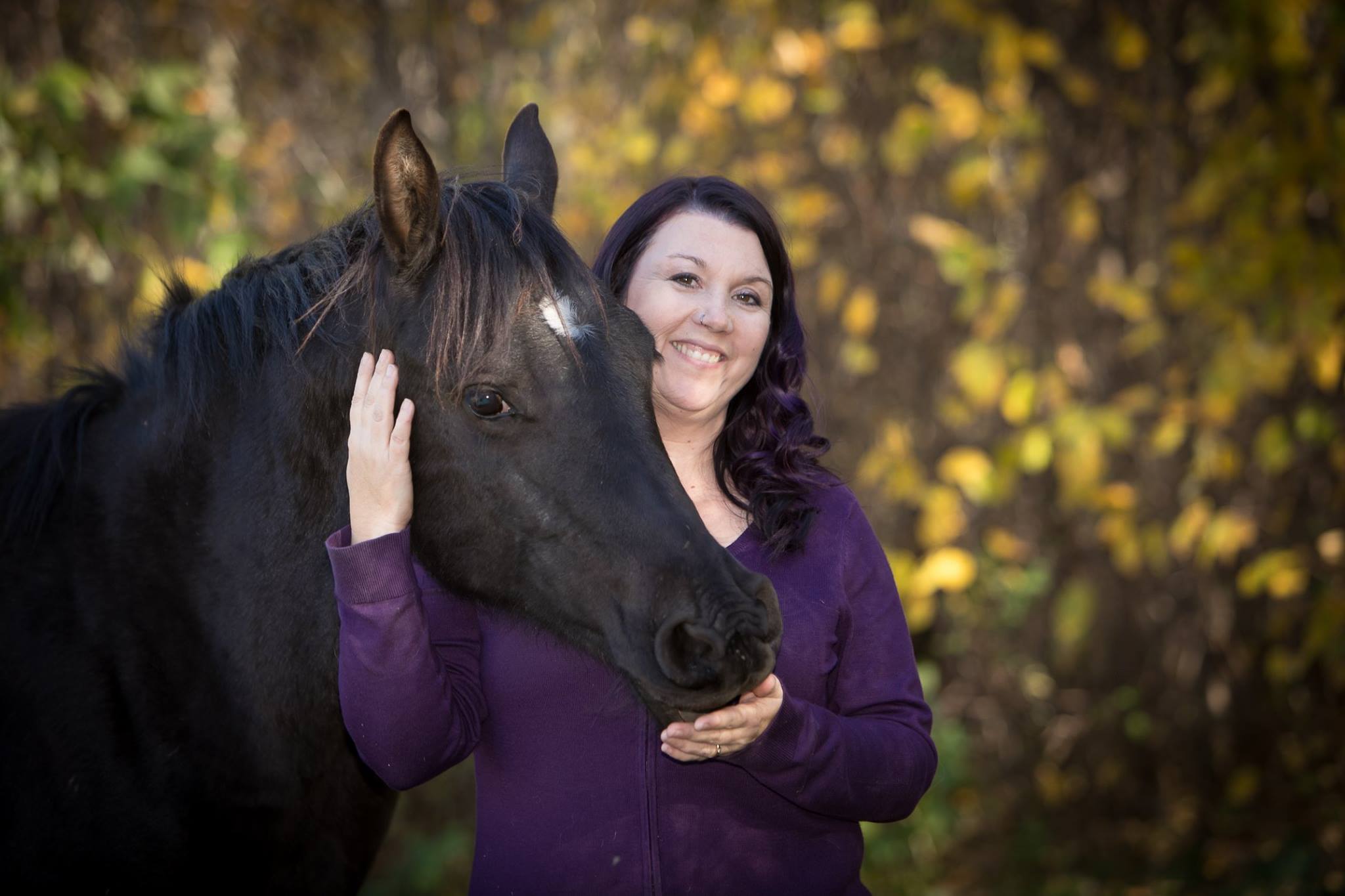 Woman smiling while gently holding her black horse in an autumn forest, capturing a joyful and intimate moment between horse and human during a photography session.